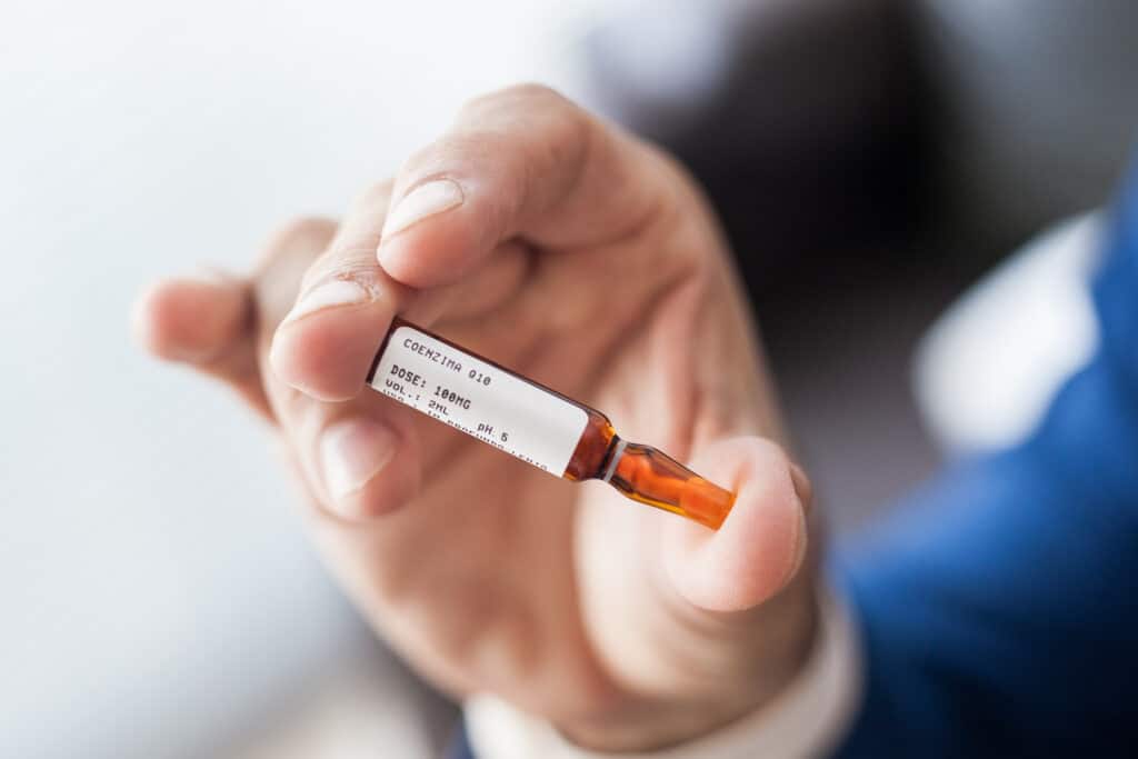 Mão de médico de terno azul segurando ampola de Coenzima Q10 100mg injetável, dose e volume visíveis no rótulo